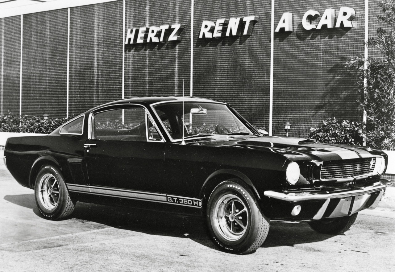 Schwarz-weißes Foto eines Shelby GT 350-H (erste Generation) vor einer Hertz-Filiale mit der Aufschrift 'HERTZ RENT A CAR'.